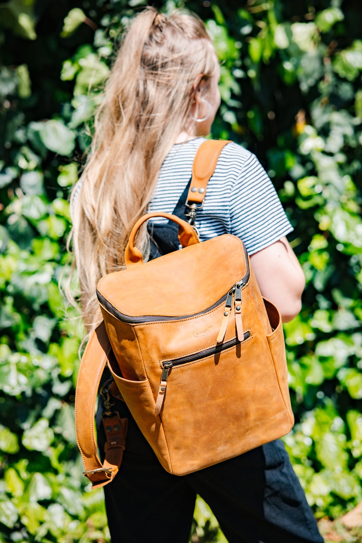 All in One Backpack Medium in Tobacco worn by a woman in a striped shirt against green foliage