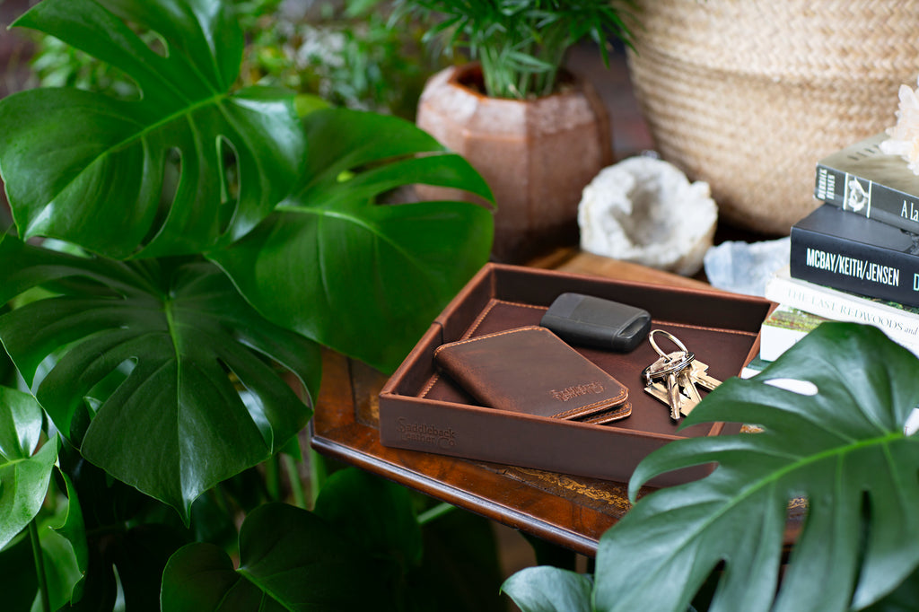 The Simple Leather Valet Tray in Dark Coffee Brown with a wallet, key fob and keys on a side table among leafy plants