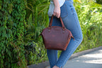 Dovetail Satchel in Chestnut, carried by its braided handles along a leafy garden path