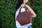Dovetail Satchel in Chestnut, held over the shoulder by its braided handles against green ivy