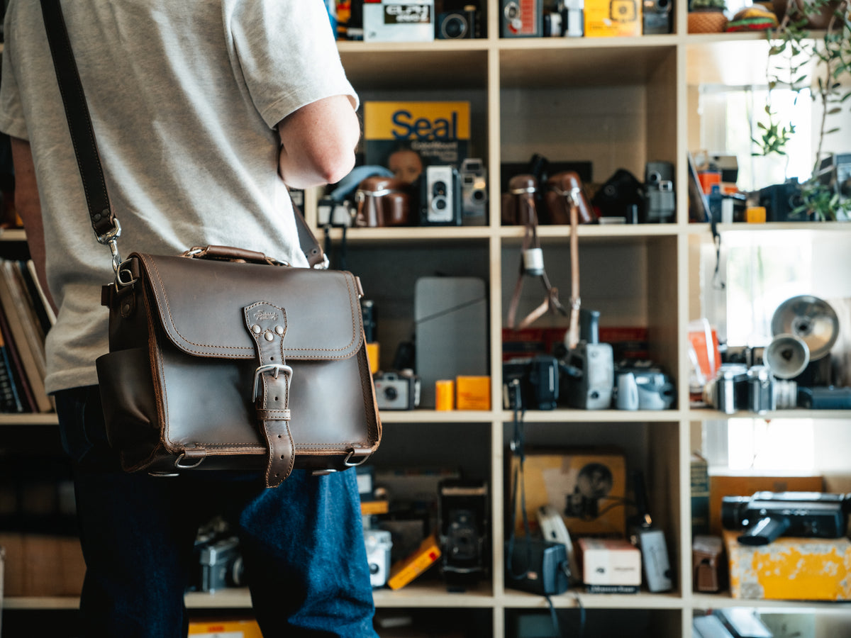 Photographer's Satchel in Dark Coffee Brown, carried by a man in front of shelves of vintage cameras