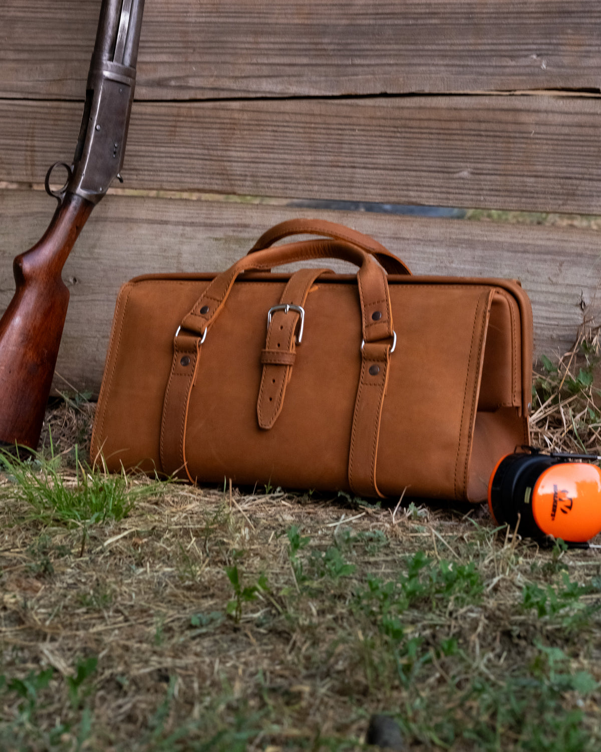Leather Range and Tool Bag in Tobacco on grass against a wooden wall beside a shotgun and orange ear muffs