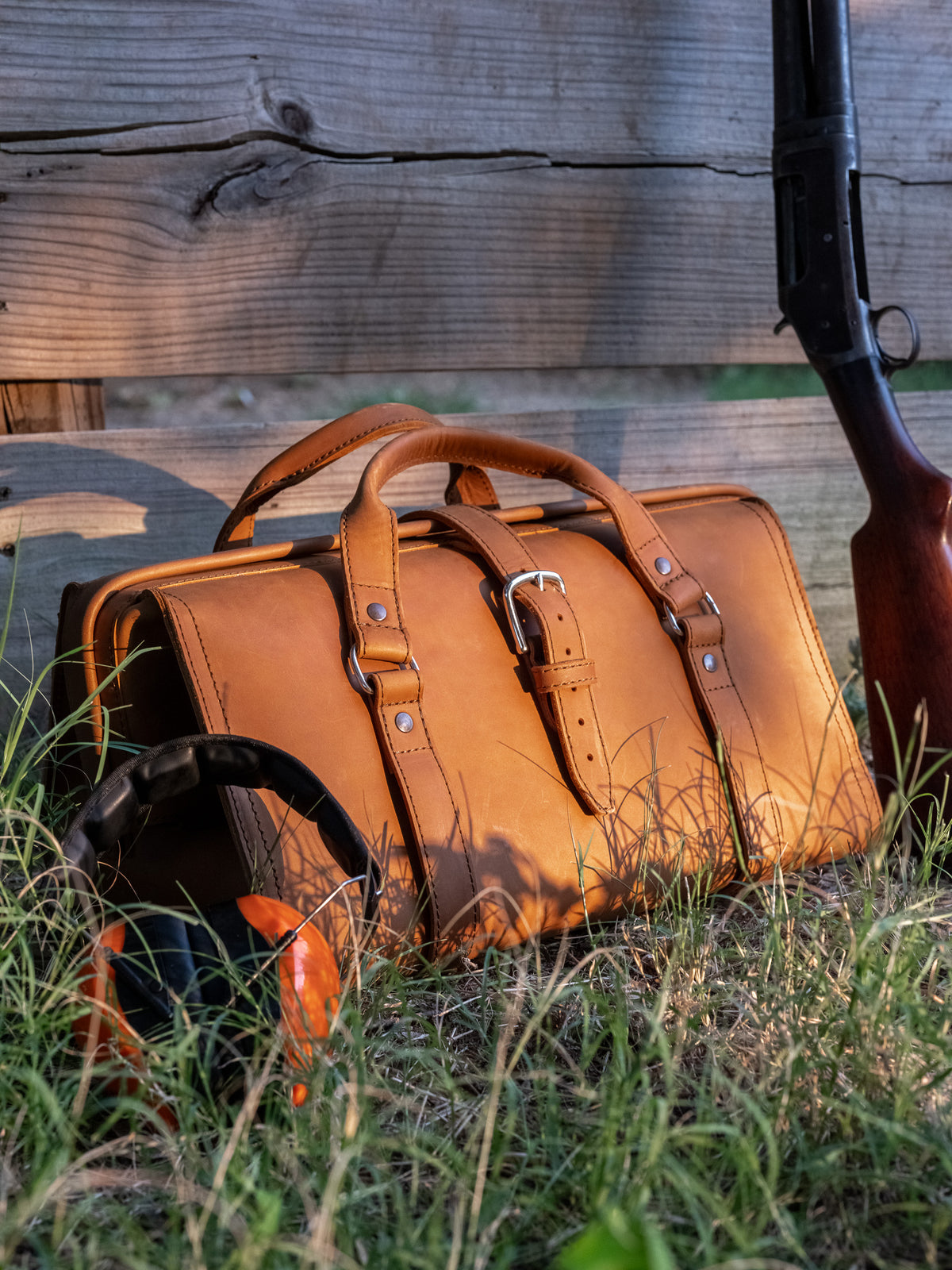 Leather Range and Tool Bag in Tobacco on tall grass by a fence with ear muffs and a shotgun leaning nearby