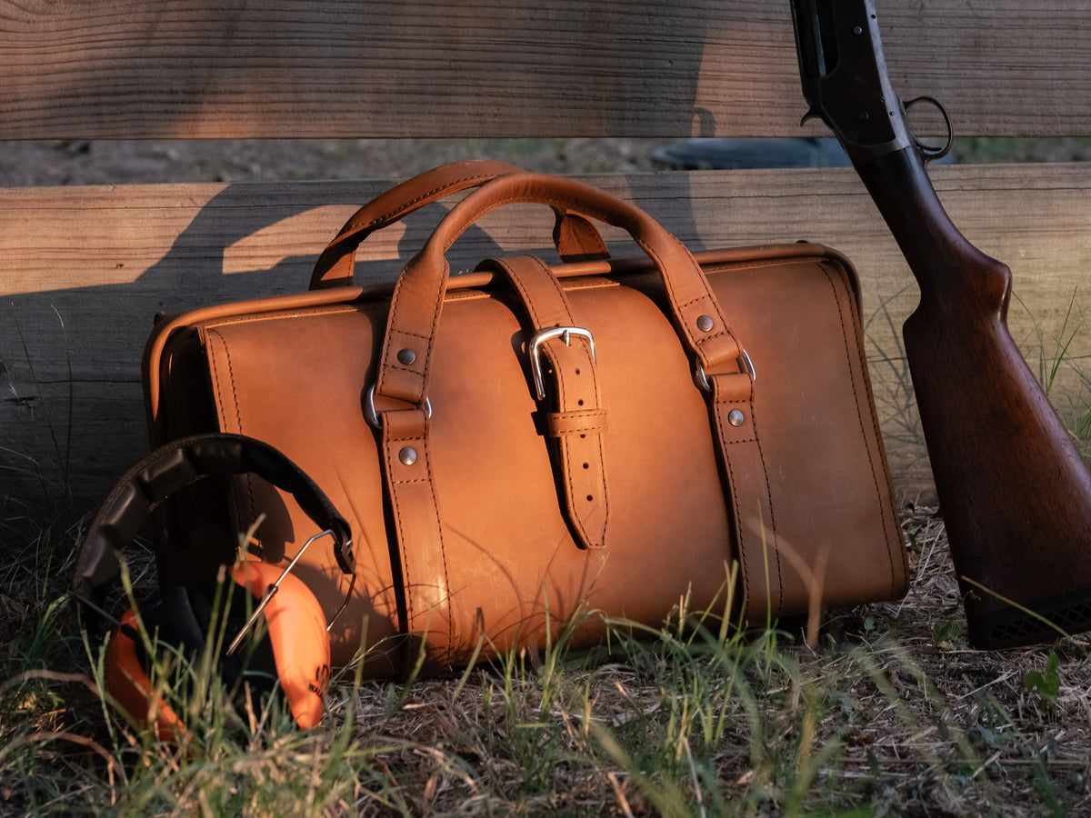 Leather Range and Tool Bag in Tobacco on grass by a wooden fence with ear muffs and a shotgun propped beside it