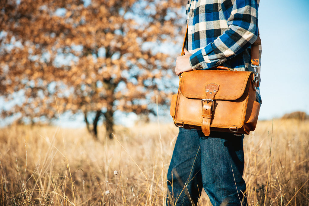 Photographer's Satchel in Tobacco, carried at the hip by a man in a plaid shirt in a dry grass field