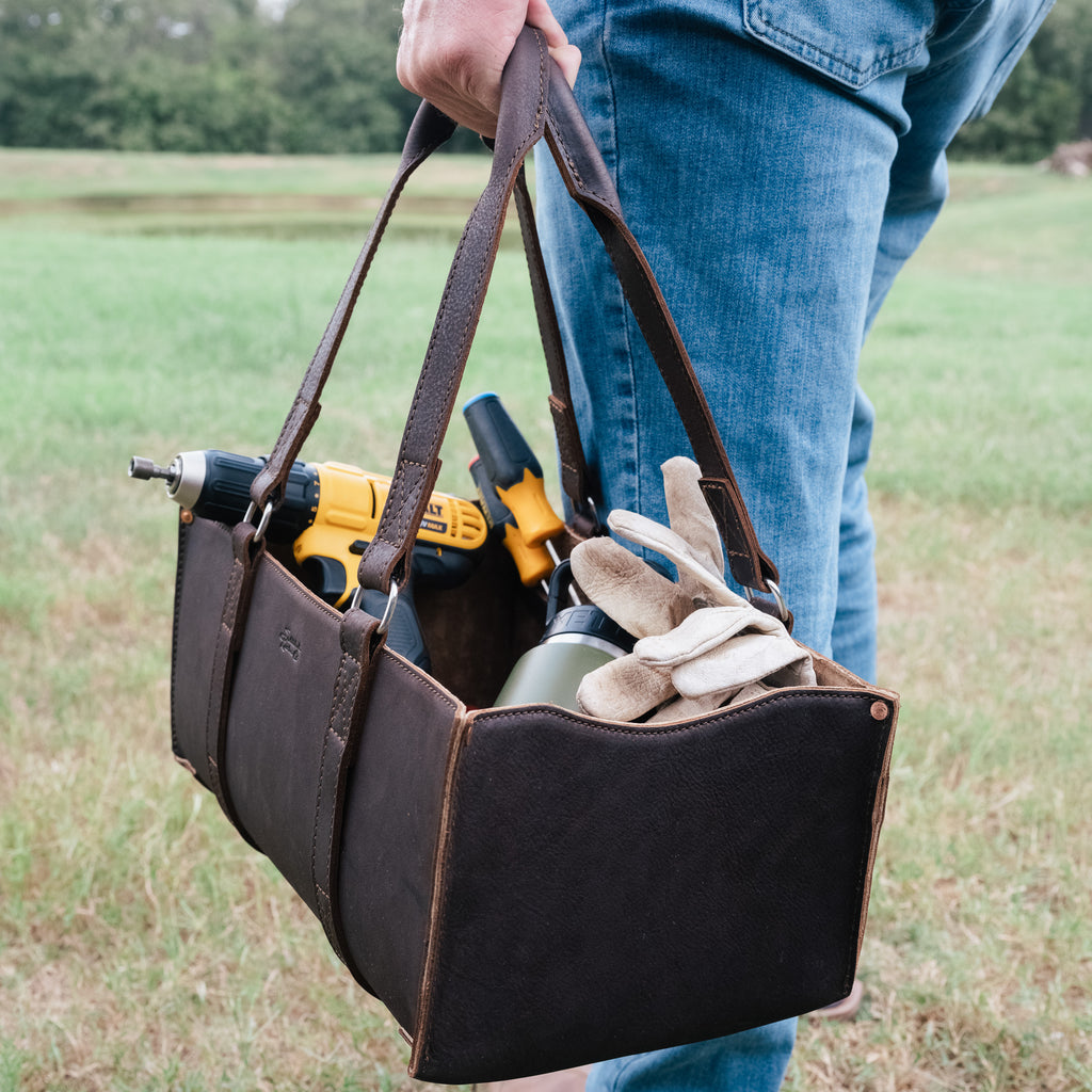 A man in jeans carrying the Range and Tool Tote in Dark Coffee Brown loaded with tools across a field