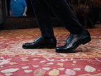 Wingtip Shoes in Raven Black, worn with dark trousers on a red patterned rug