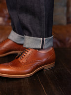 Wingtip Shoes in Natural Tan, worn with cuffed selvedge jeans on a dark wood floor