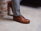 Wingtip Shoes in Natural Tan, worn while leaning against a brick wall, one foot raised