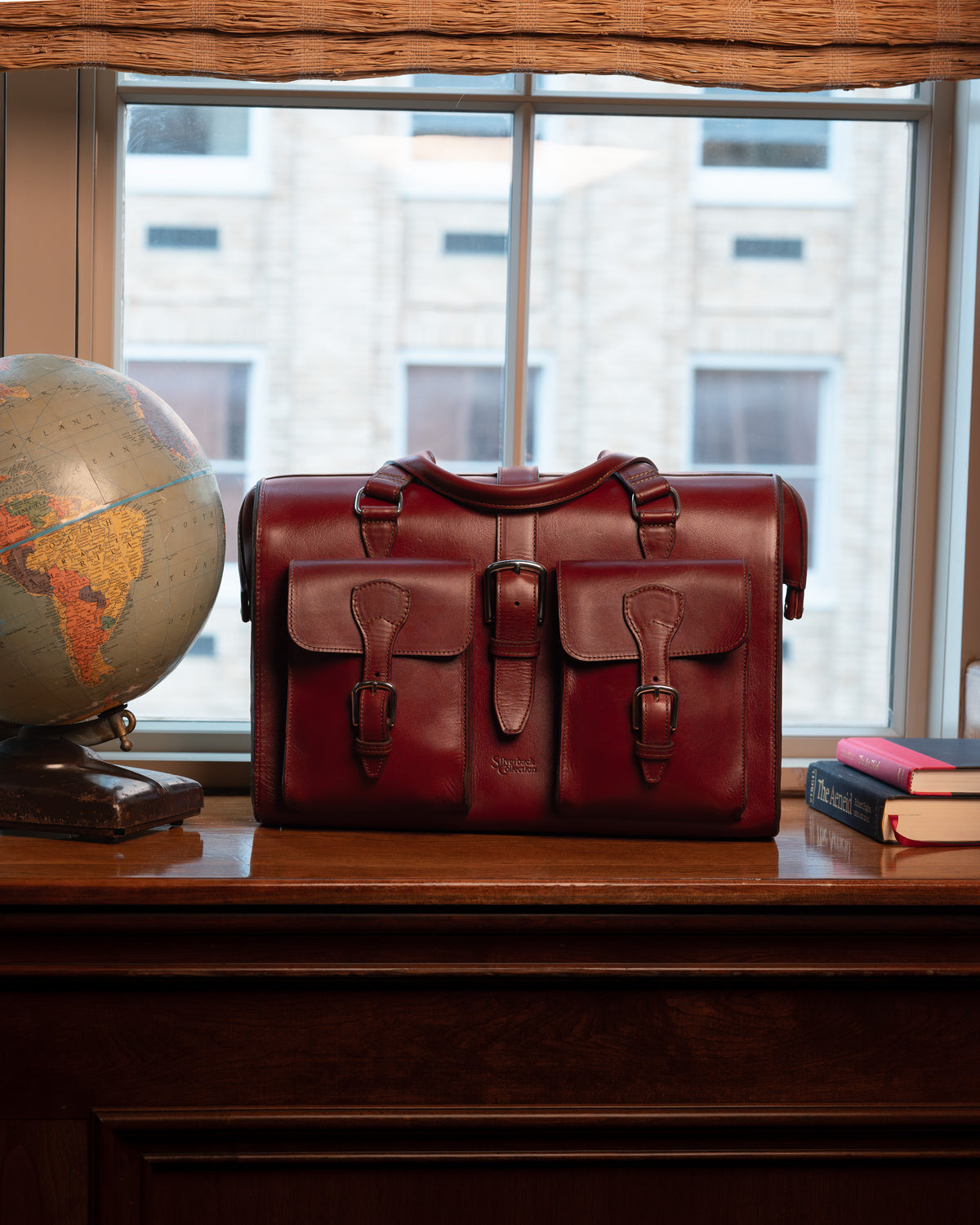 Blackstone Barrister Briefcase in Oxblood on a wooden sill by a window, between a globe and stacked books