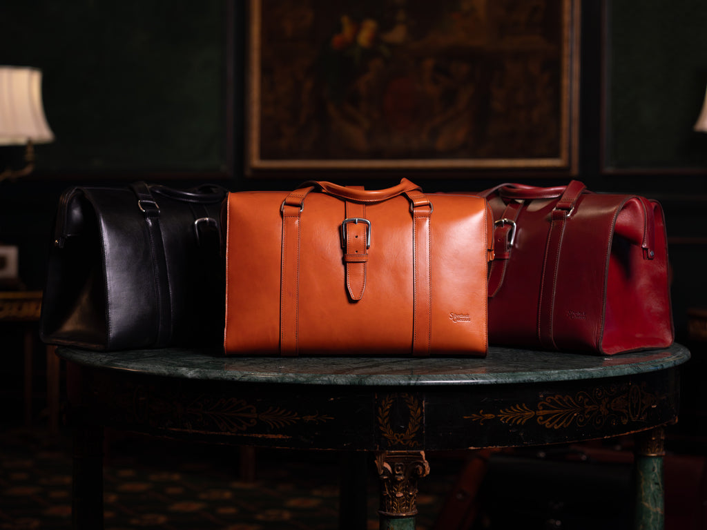 Taylor Duffle Bag in Raven Black, English Tan and Oxblood side by side on a marble table