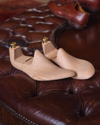 Shoe Tree pair, side view on a tufted leather ottoman with brown shoes behind