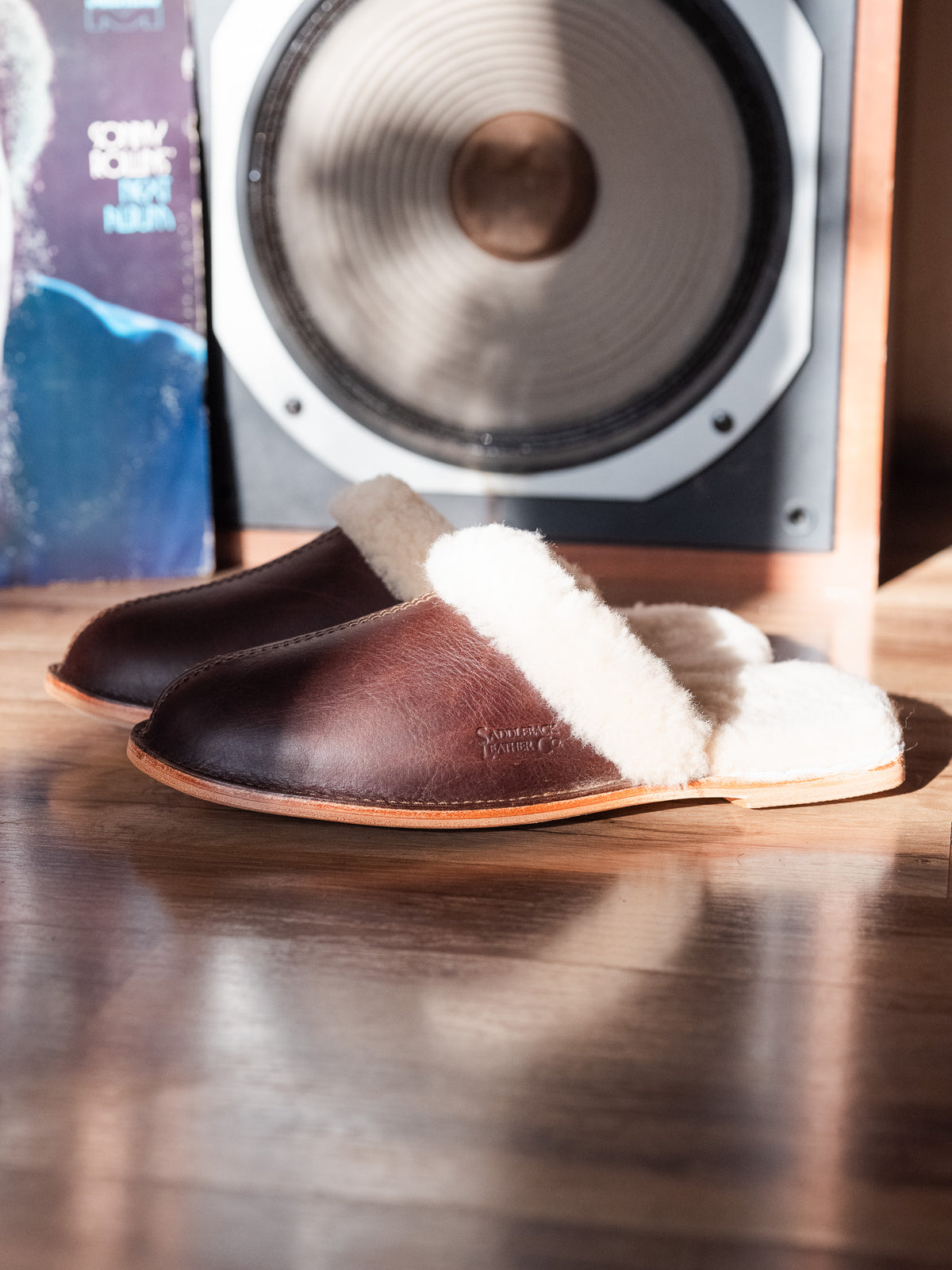 Men's Sheepskin Slippers in Dark Coffee Brown, side view of pair on a wood floor in front of a speaker