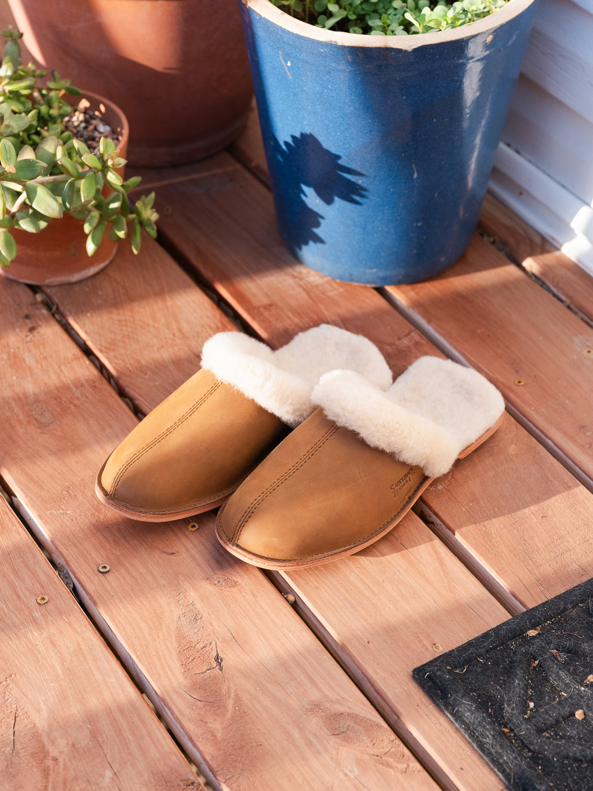 Men's Sheepskin Slippers in Tobacco, pair on a sunny wooden deck by potted plants