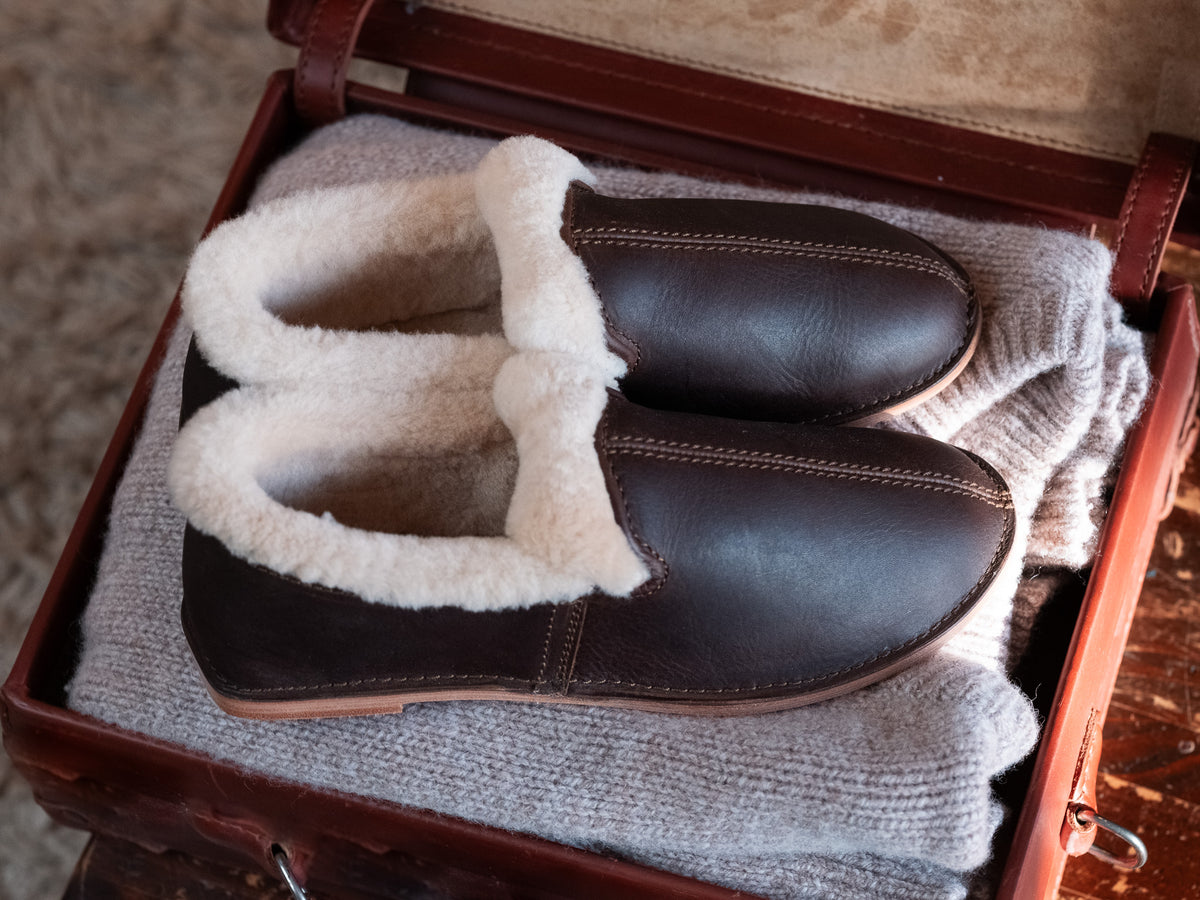 Pair of Sheepskin Moccasins in Dark Coffee Brown resting on a folded gray sweater inside an open leather case