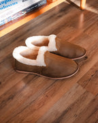 Pair of Sheepskin Moccasins in Tobacco with cream shearling cuffs on a wood floor beside a low bookshelf