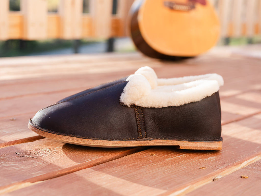 Sheepskin Moccasin in Dark Coffee Brown with shearling cuff, side view on a wooden deck with a guitar behind