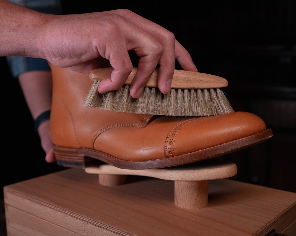 Luxury Shoe Shine Kit in use, a hand brushing a tan leather cap-toe boot on the wooden shine stand
