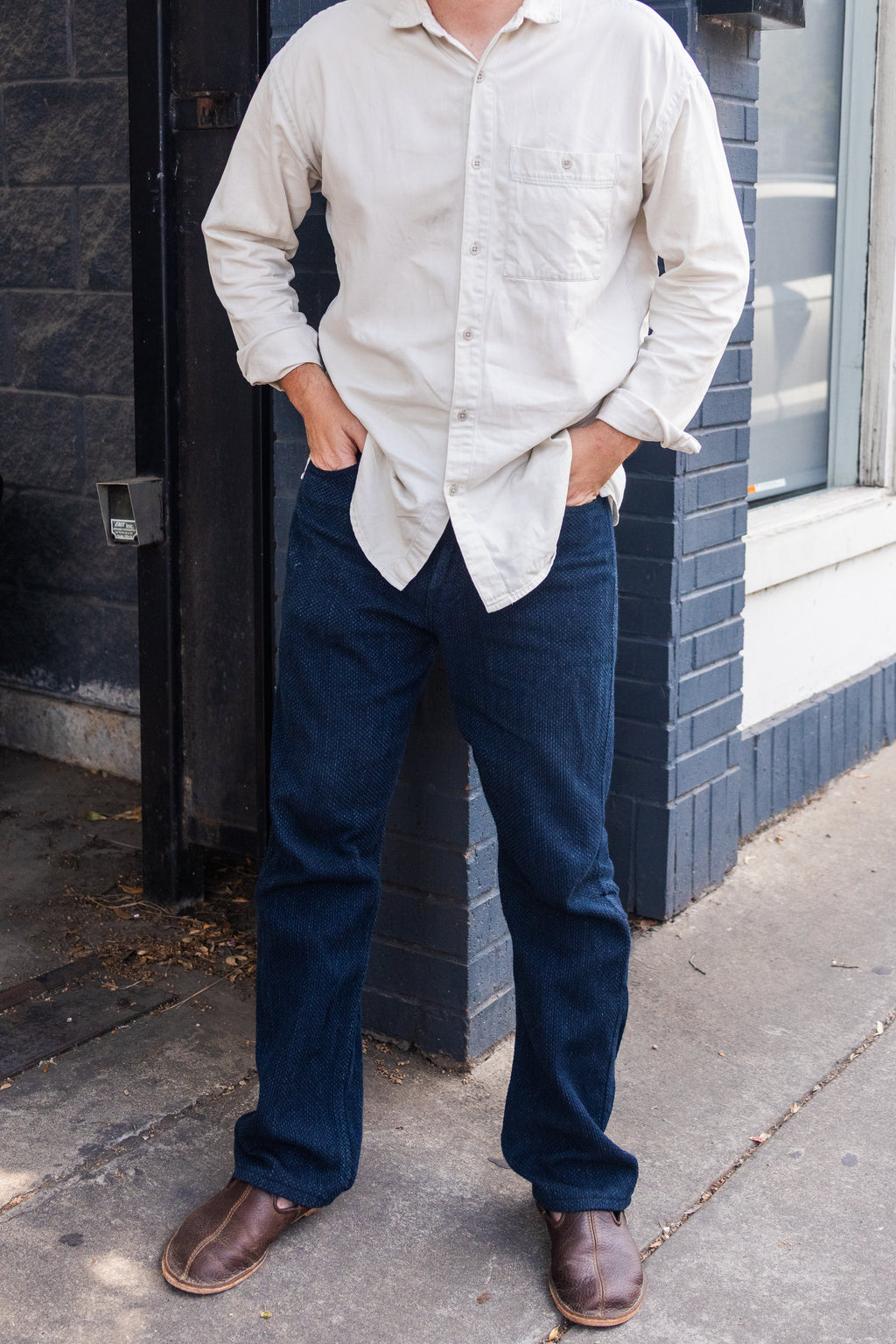 A man in jeans standing by a brick wall wearing Barefoot Slip-on Mules in Dark Coffee Brown