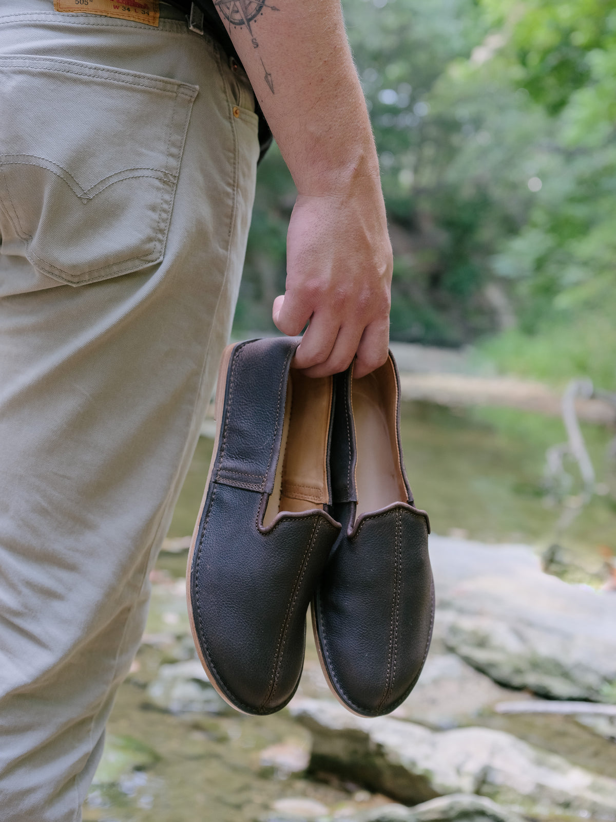 A man carrying a pair of Barefoot Slip-on Shoes in Dark Coffee Brown beside a rocky creek