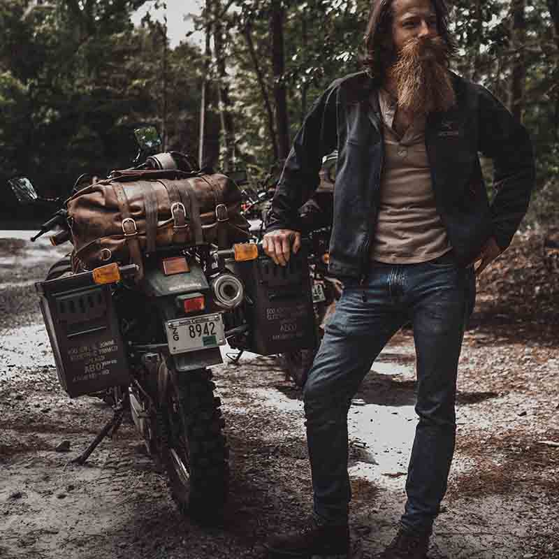 24" Waterbag in brown, strapped to a loaded adventure motorcycle beside its rider on a forest trail