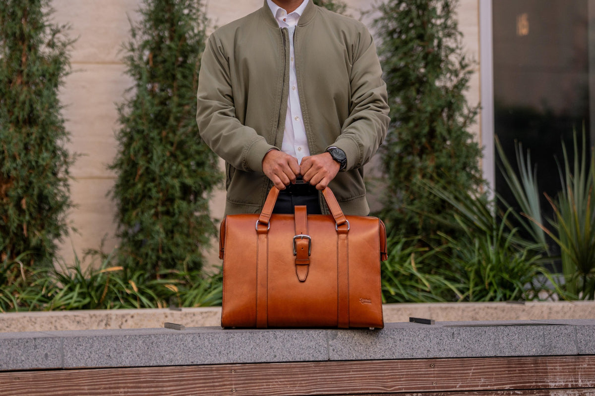 Man in an olive bomber jacket holding a tan leather doctor's duffle by its handles outdoors
