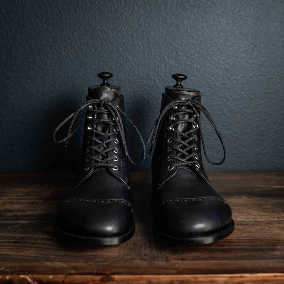 Officer's Boot in Raven Black, front view of the pair with shoe trees on a wooden table