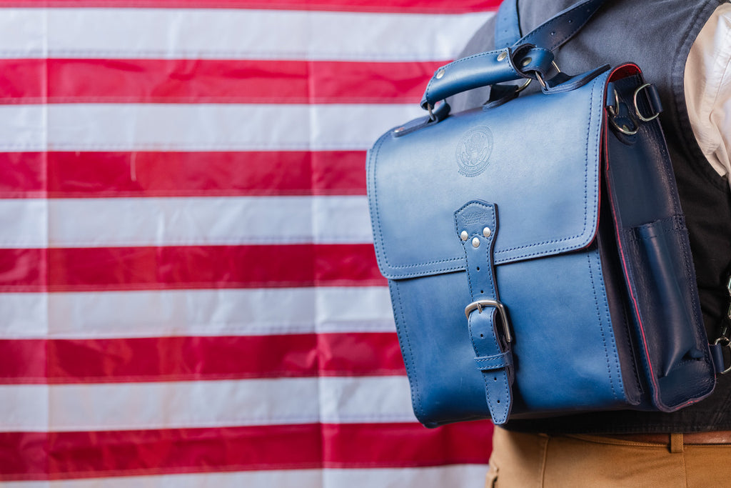 Saddleback Club Membership Paul Revere Leather Messenger Bag Backpack in Red, White and Blue, worn in front of an American flag