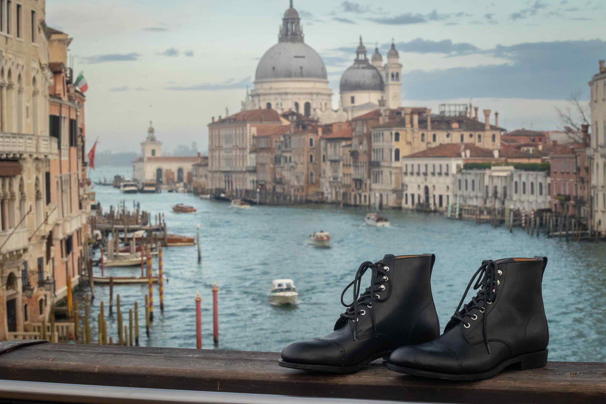 A pair of Raven Black Officer's Boots on a railing above Venice's Grand Canal, the domes of Santa Maria della Salute behind them.