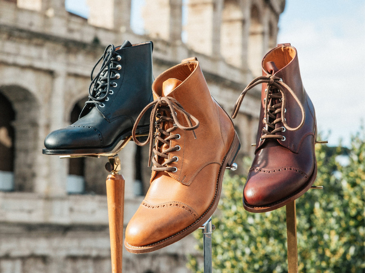 Three Officer's Boots — Raven Black, English Tan and Oxblood — raised on boot stands in front of the Colosseum.