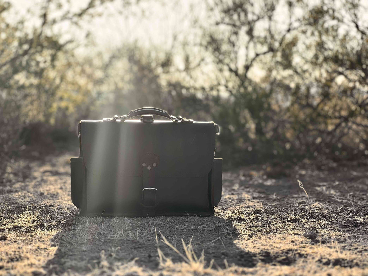 Full grain leather slim briefcase standing on a dirt trail, backlit by the sun through desert brush