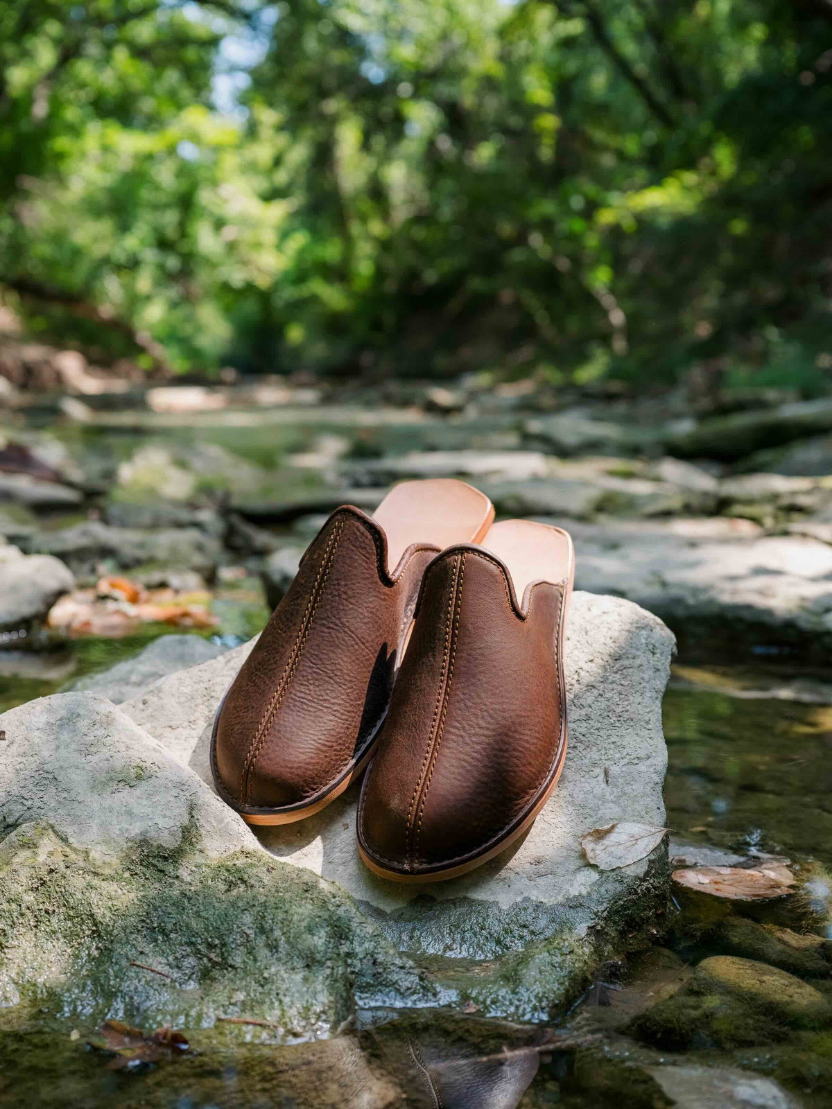 Pair of brown leather barefoot mule slip-on shoes on a rock in a wooded creek