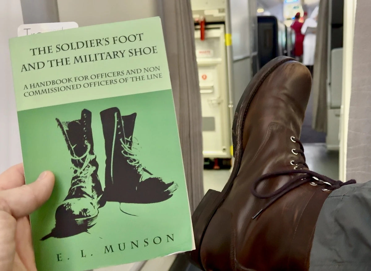 Hand holding the book The Soldier's Foot and the Military Shoe beside a brown leather boot on an airplane