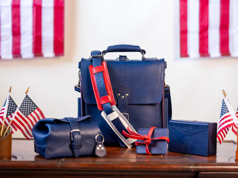 The whole Fourth Edition bundle laid out on a desk with small United States flags: the messenger bag, toiletry bag, cash box, coaster set and keychain, all in navy leather