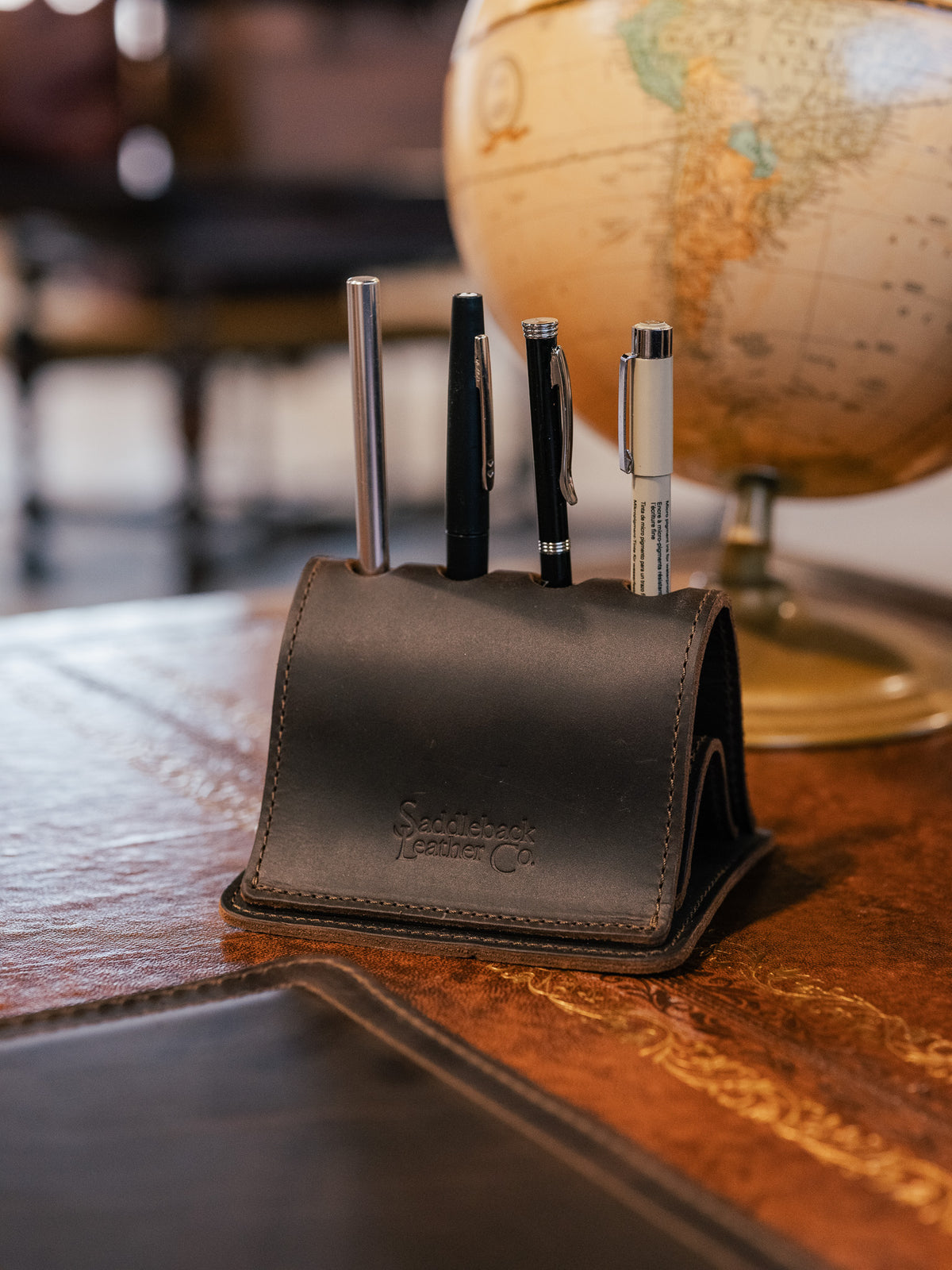 Pen Holder Bell Curve in Dark Coffee Brown leather holding four pens on a wooden desk with a globe behind