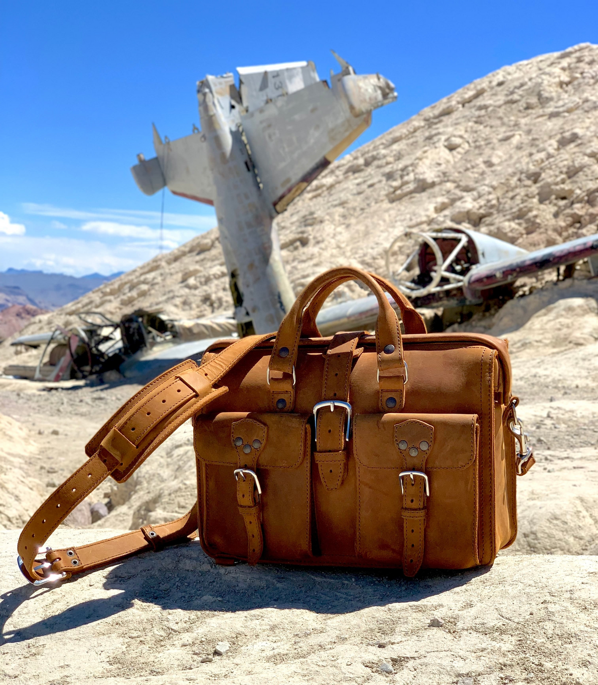 Tan Saddleback Leather Flight Bag pilot case on a rocky desert slope with a crashed airplane behind it