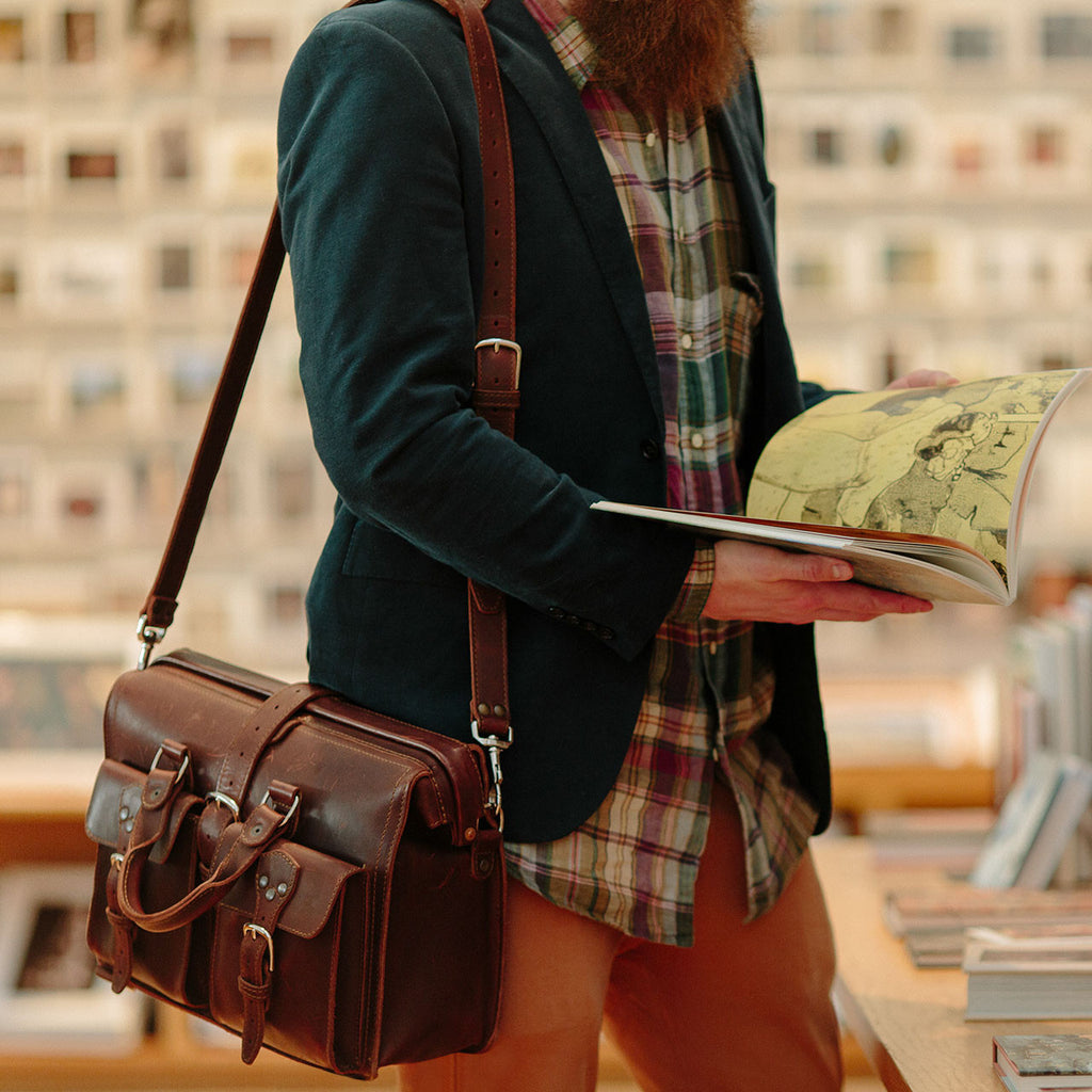 Man in a bookshop reading a book while carrying the Flight Bag Leather Briefcase in chestnut on its shoulder strap