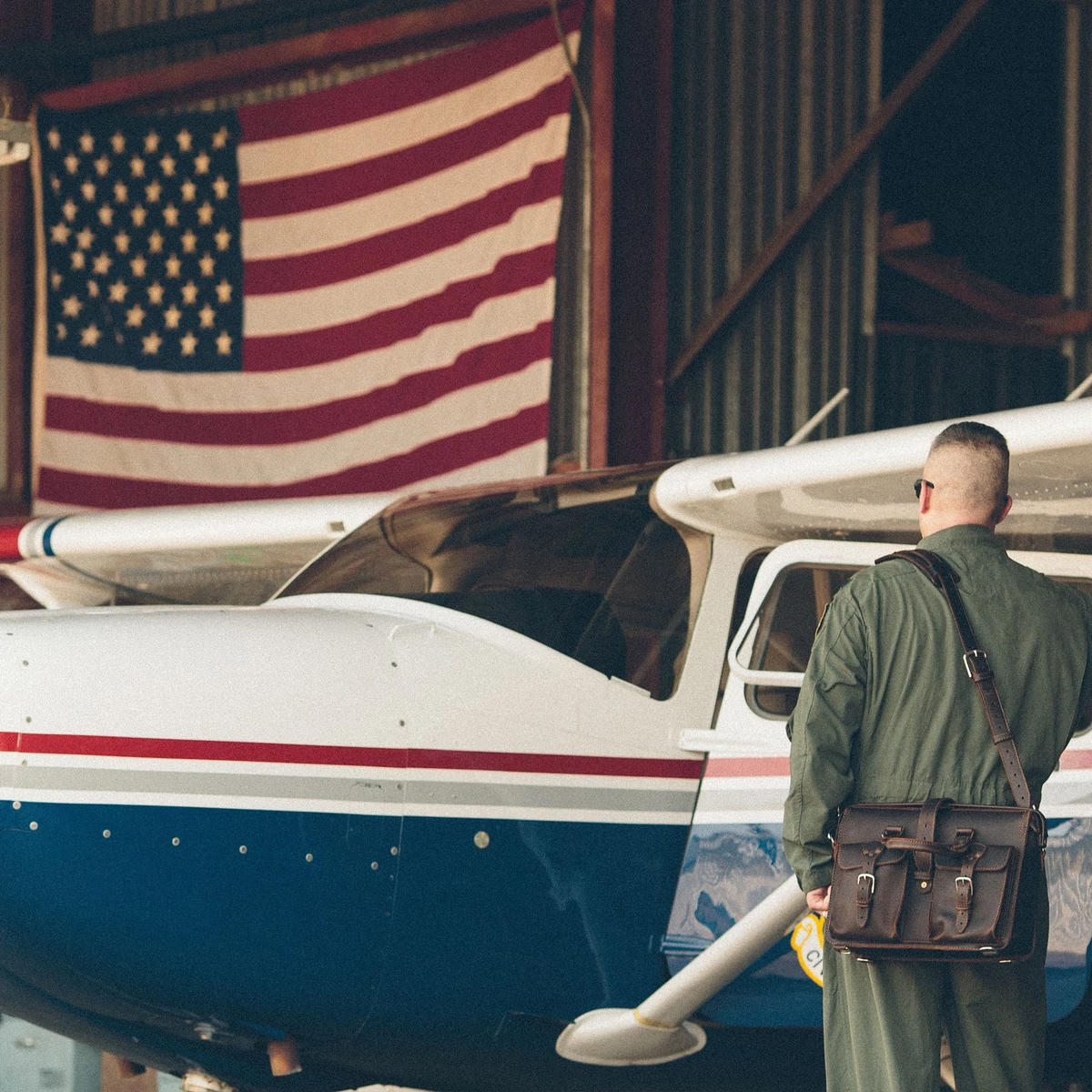 Pilot in an olive flight suit carrying a dark brown leather Flight Bag beside a small plane in a hangar with an American flag