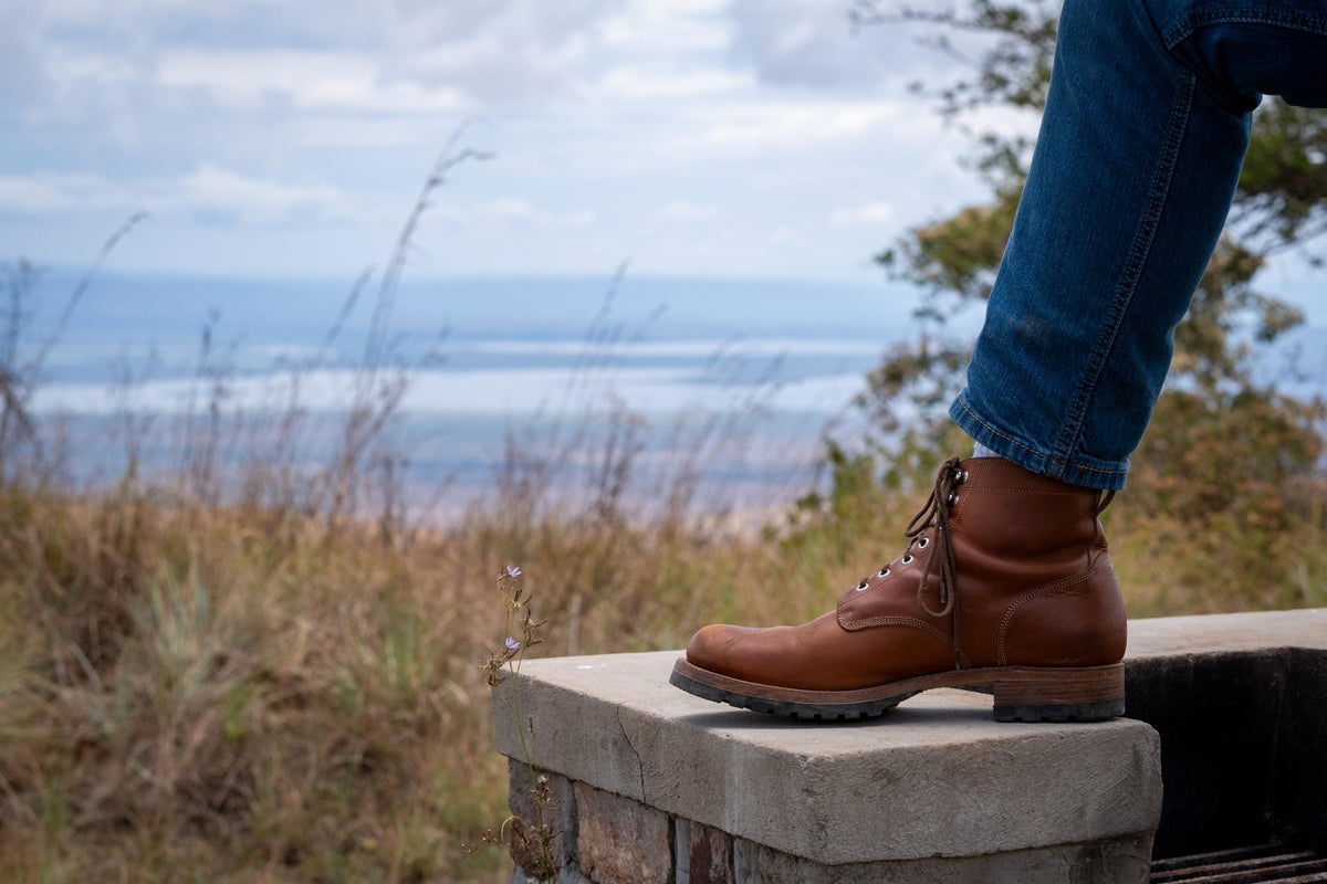 Huntsman Boots in brown leather, one boot on a stone post overlooking a grassy field and water
