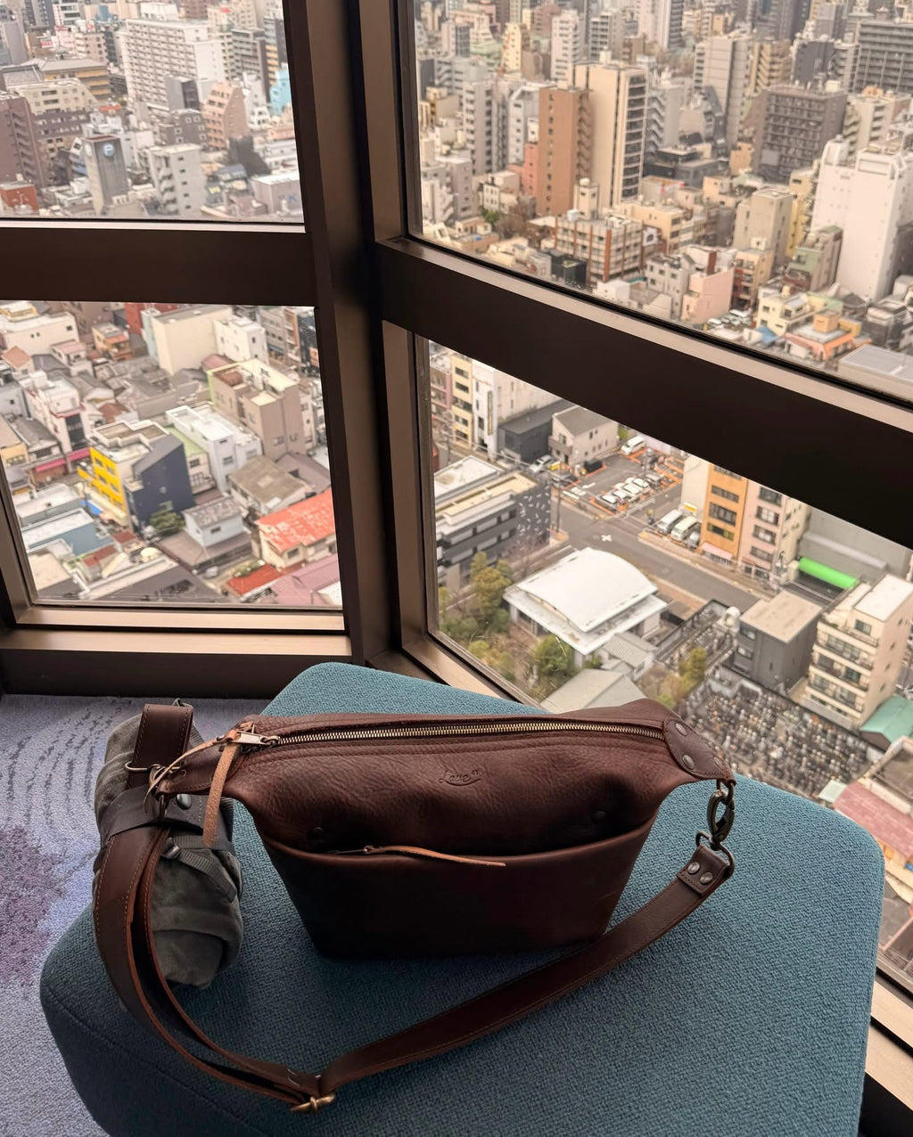 London Sling Small in Dark Coffee Brown, resting on a blue chair by a high-rise window with a city view