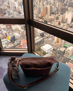 London Sling Small in Dark Coffee Brown, resting on a blue chair by a high-rise window with a city view