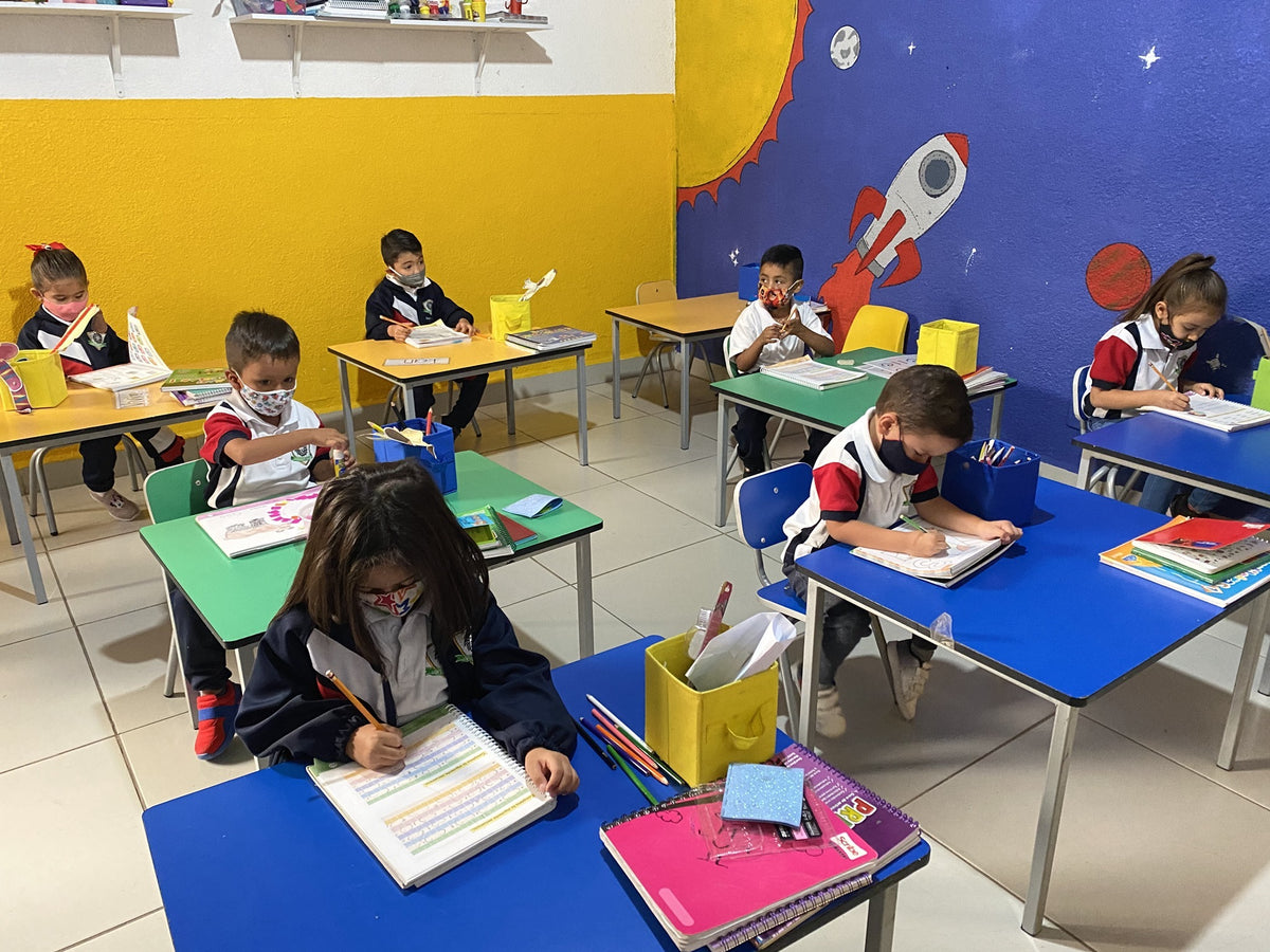 Young children in school uniforms and face masks working at brightly coloured desks in a classroom at Lion's Heart Academy