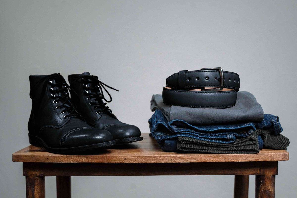 Italian Casual Belt in Raven Black, coiled on folded jeans beside black leather boots