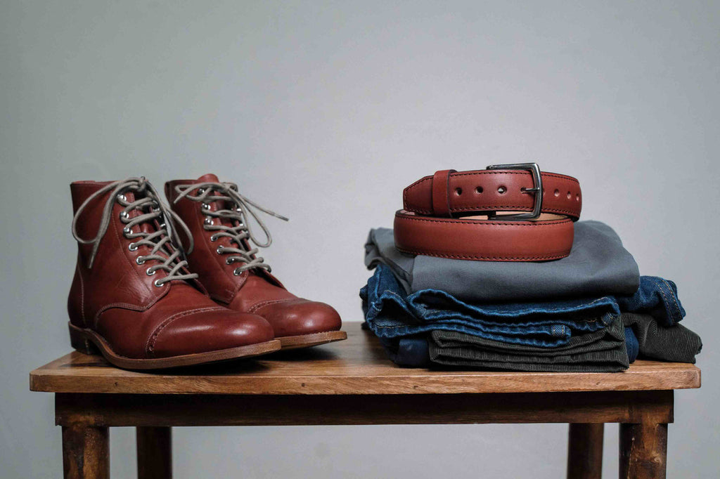 Officer Boot in Oxblood, pair on a wooden bench beside a coiled belt and folded jeans