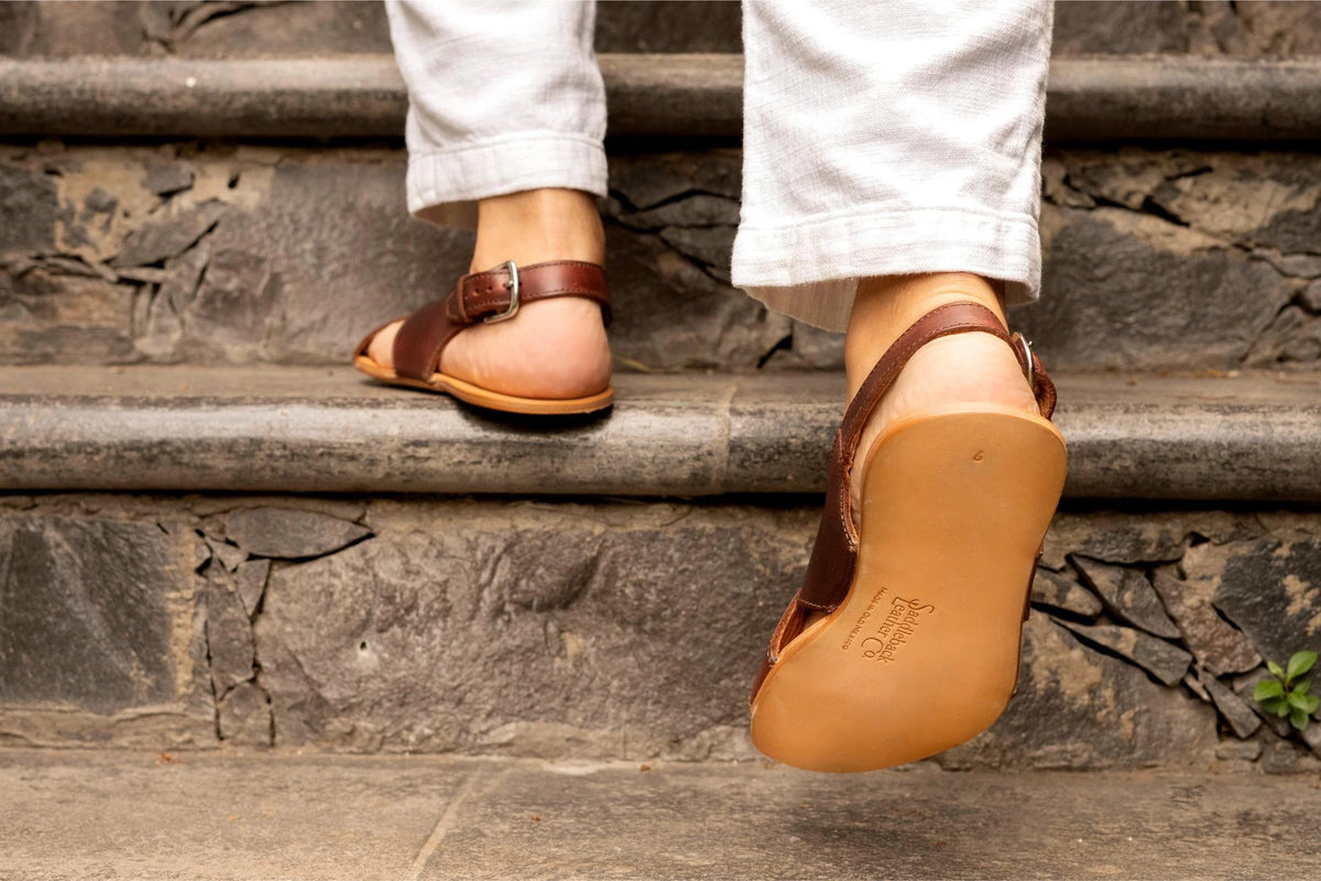 Jesus Sandals in Chestnut, back view mid-step on stone stairs showing the logo-stamped leather outsole