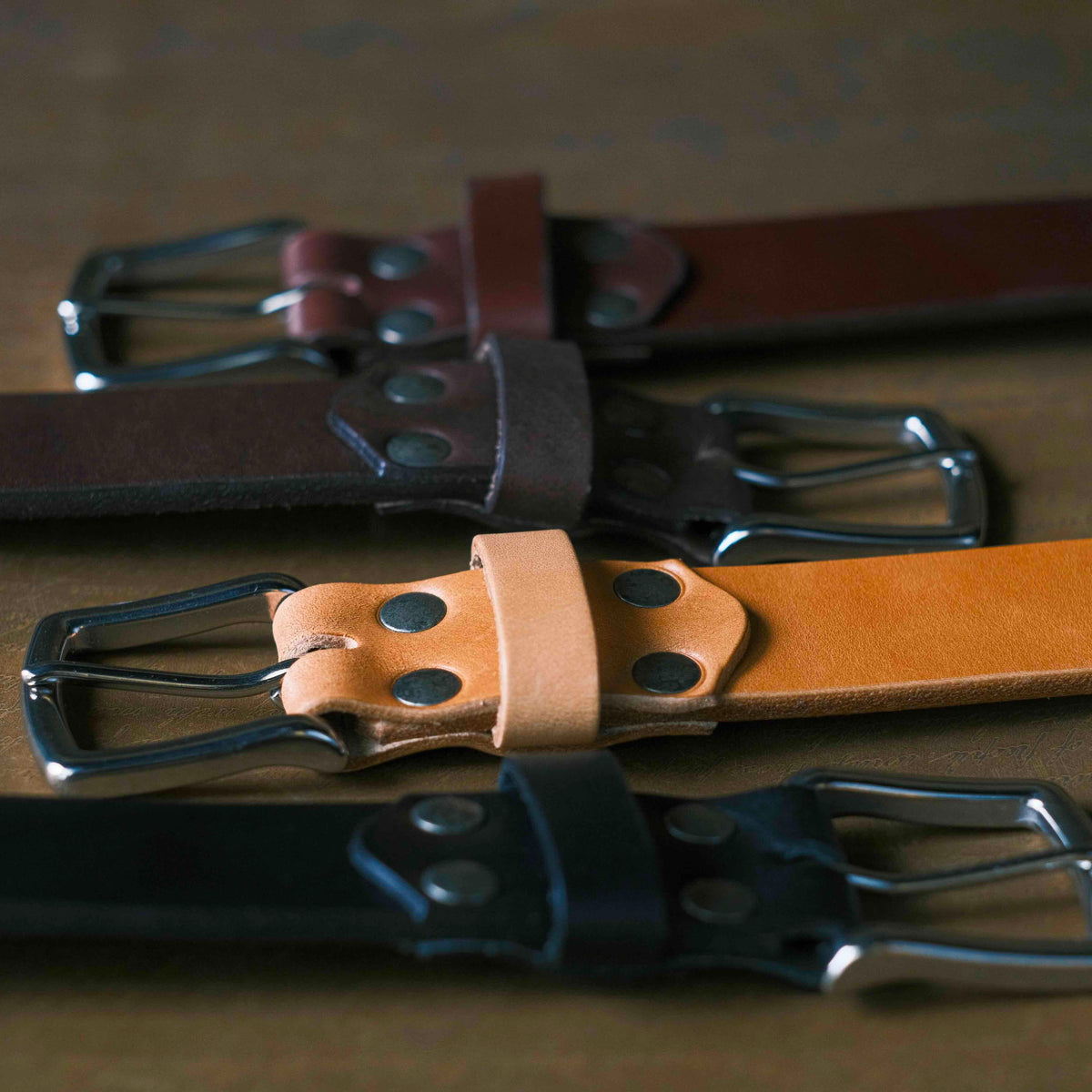 Old Bull Leather Belts in Chestnut, Dark Coffee Brown, Natural and Black laid side by side on a wood table