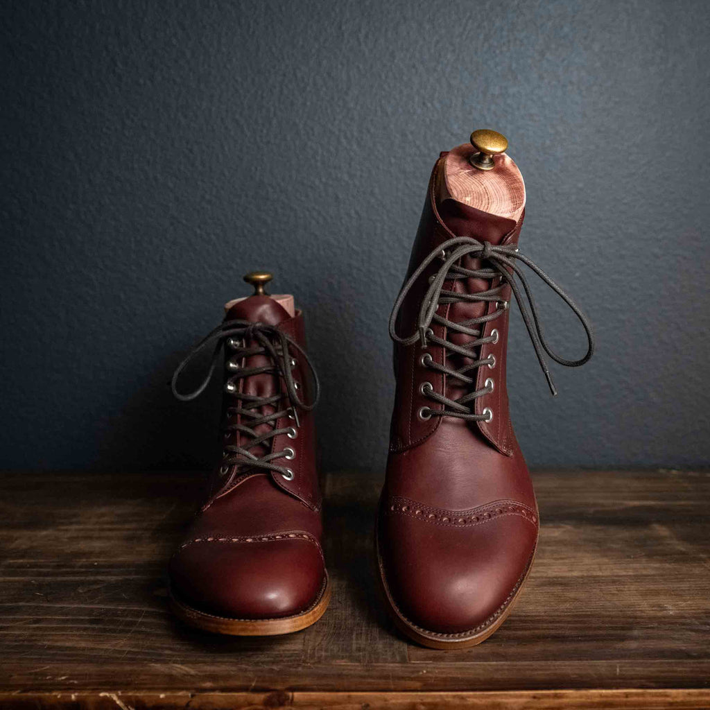 Officer Boot in Oxblood, staggered pair with dark laces on a wooden table