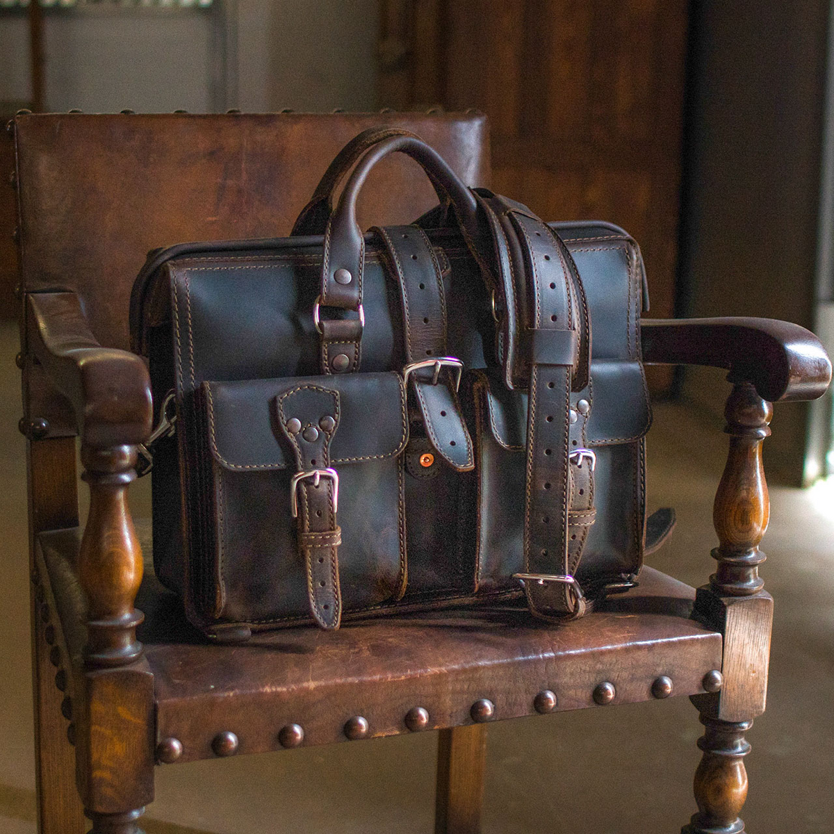 Dark brown full grain leather briefcase with buckle straps resting on an antique wooden chair