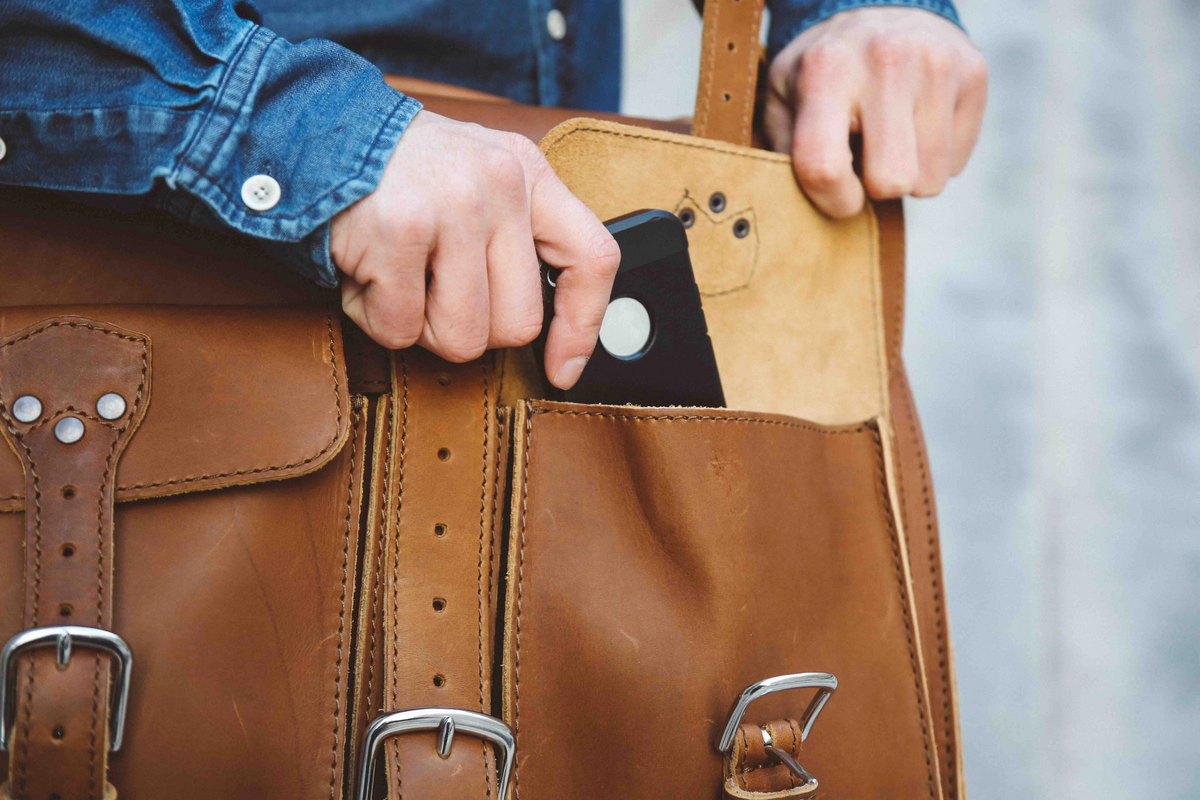 Close-up of hands sliding a phone into the front pocket of a brown leather briefcase with buckle straps