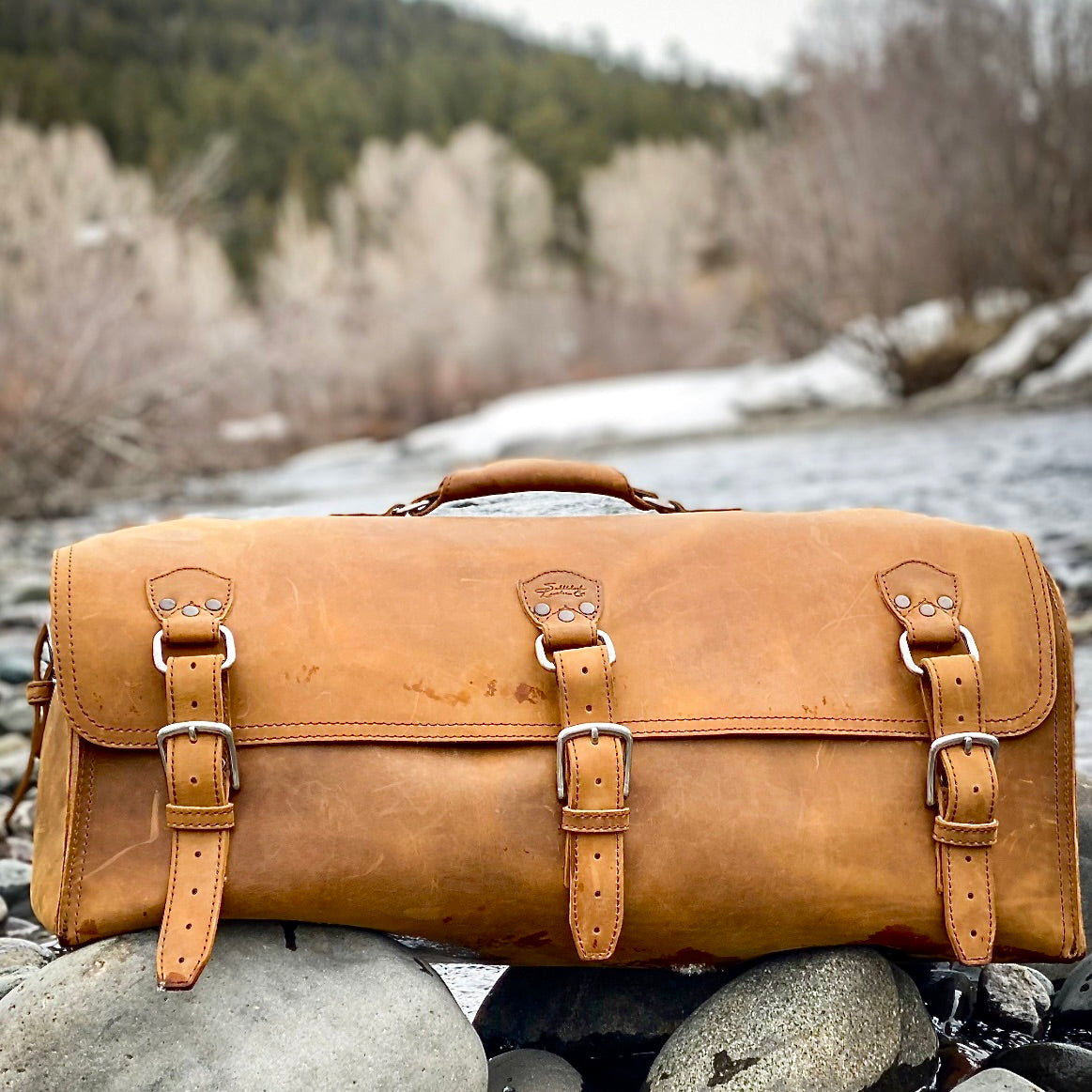 27" Adolescent Beast Duffle Bag in Tobacco resting on river rocks beside a snowy mountain stream